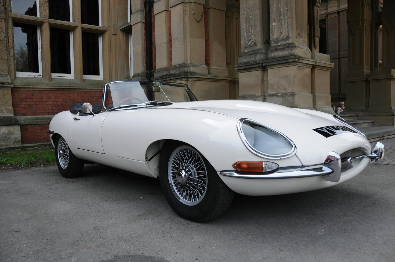 A white Jaguar E-Type Series 1 roadster parked beside a Victorian stone building