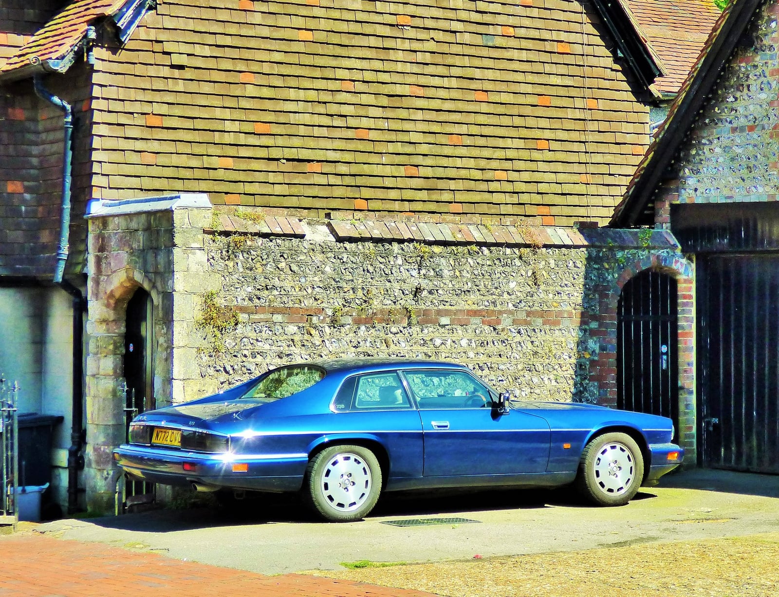 A blue Jaguar XJS coupe parked beside a flint barn, side three-quarter view