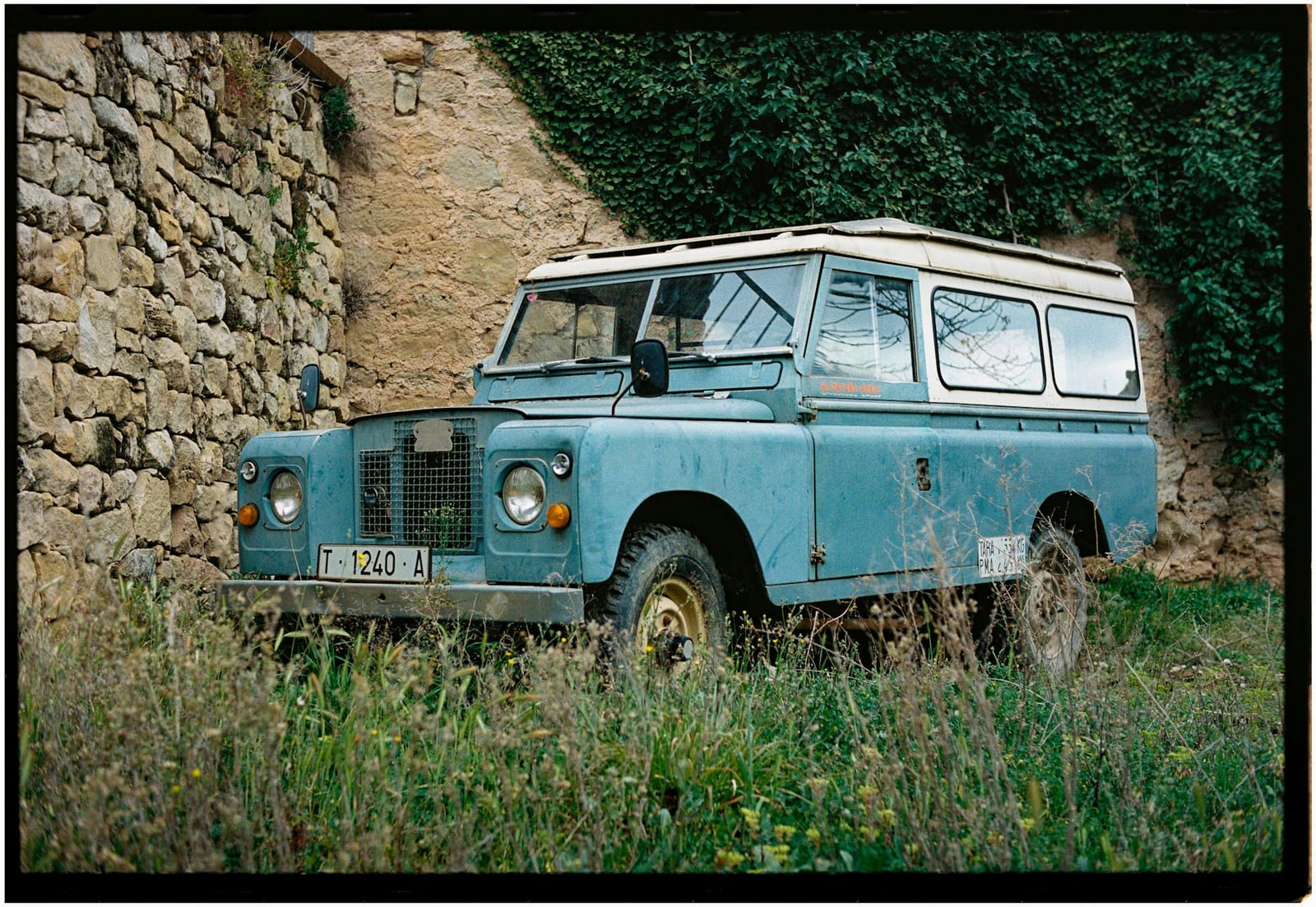 A patinated blue Land Rover Series long-wheelbase station wagon parked on grass beside an old stone wall