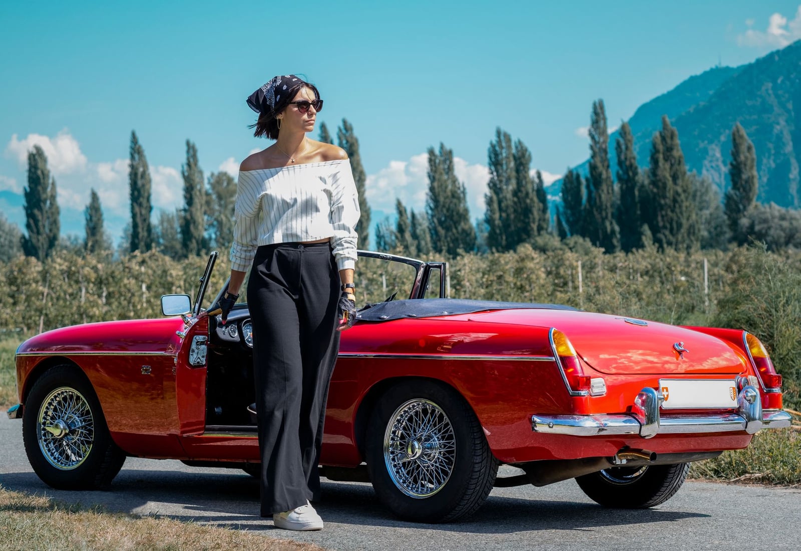 A red MG MGB roadster with chrome wire wheels parked on a street, a person standing alongside