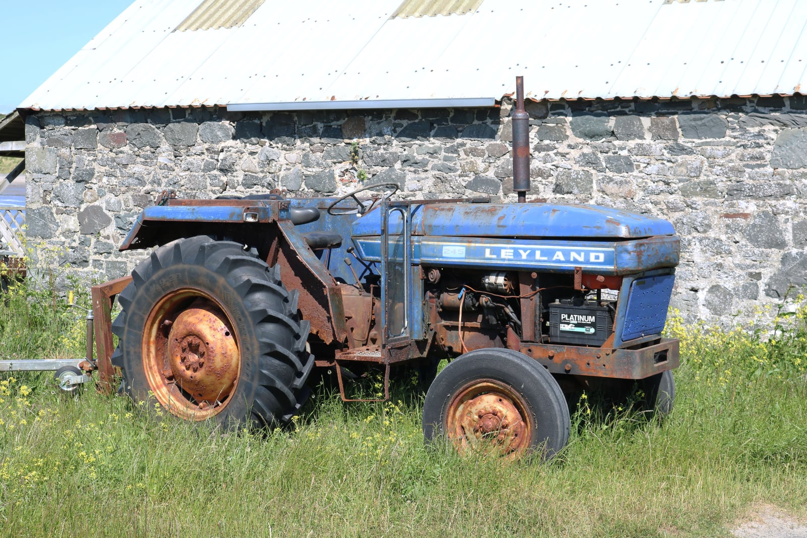 A faded blue Leyland tractor with rusty wheels standing in long grass beside a stone barn