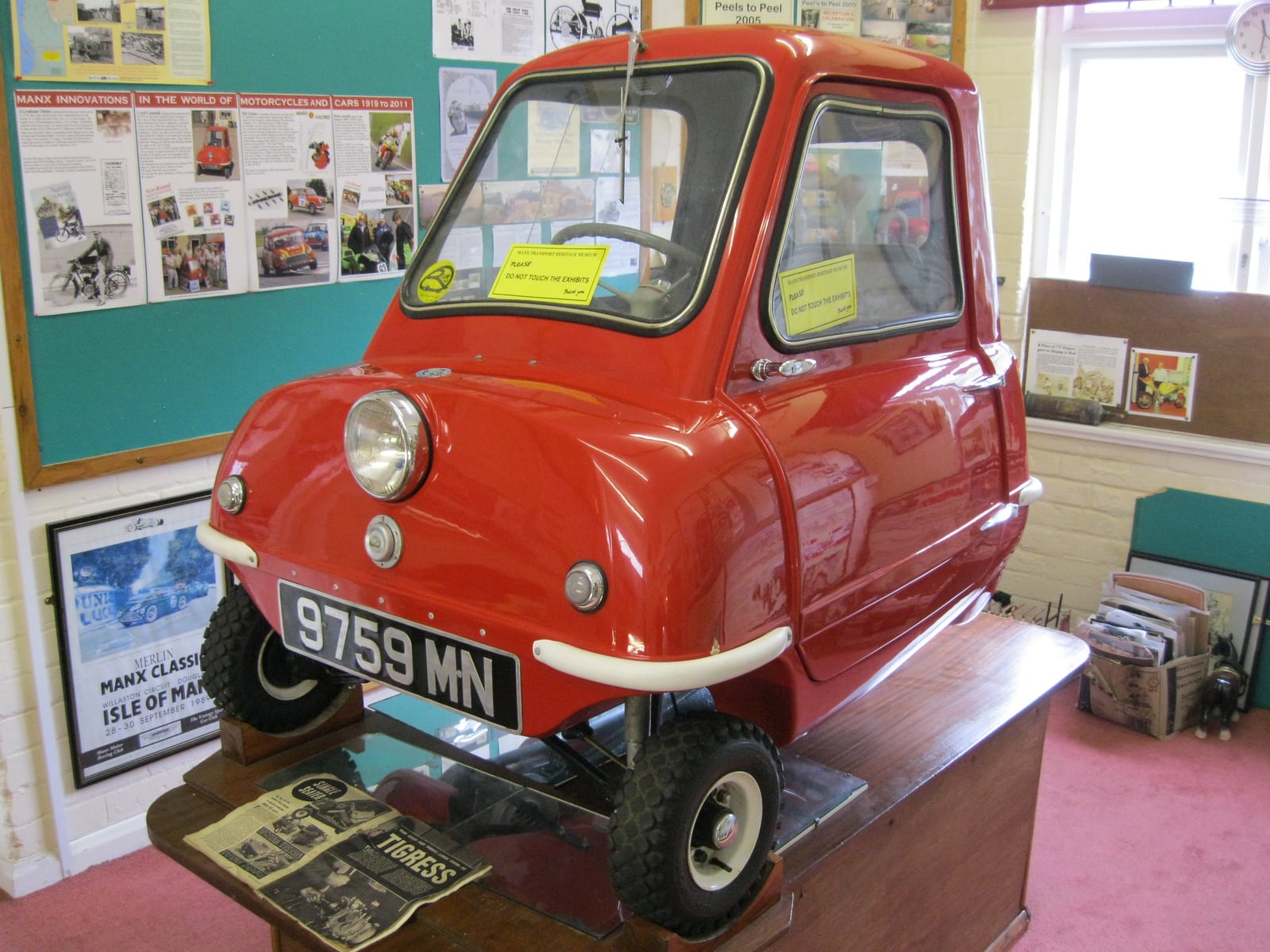 A red Peel P50 microcar on a display plinth at the Manx Transport Museum in Peel, Isle of Man, front three-quarter view showing the single door and three wheels