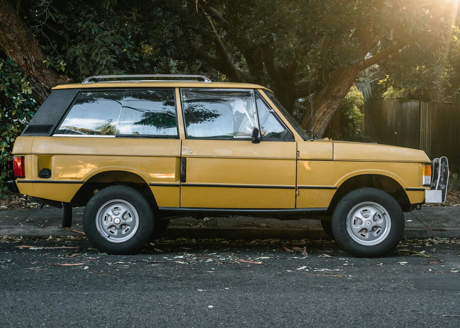 A yellow two-door Range Rover Classic, side profile, parked on a tree-lined street