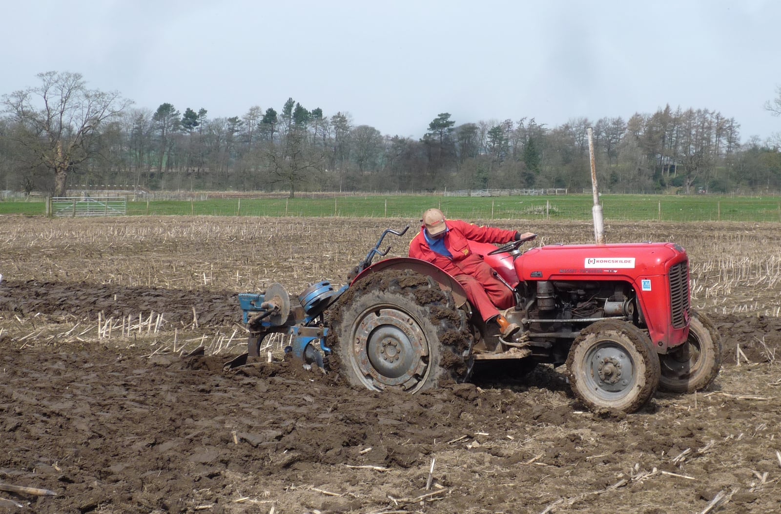 A red Ferguson 35 ploughing a field with a mounted plough, a driver aboard, fresh furrows in the foreground