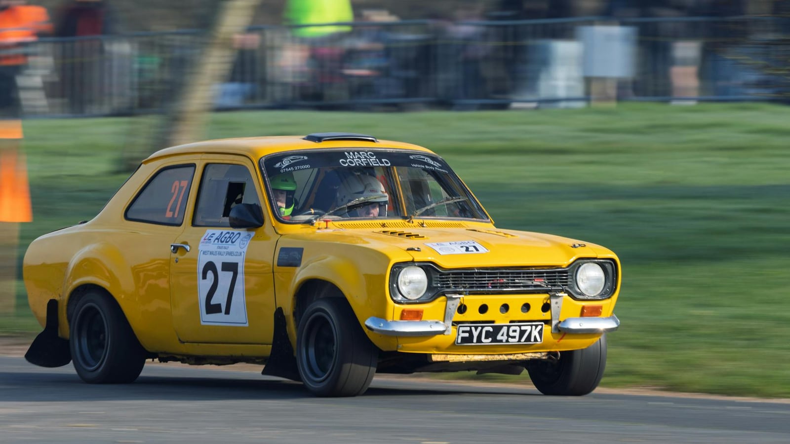 A yellow Mk1 Ford Escort rally car, number 27, cornering hard on a rally stage